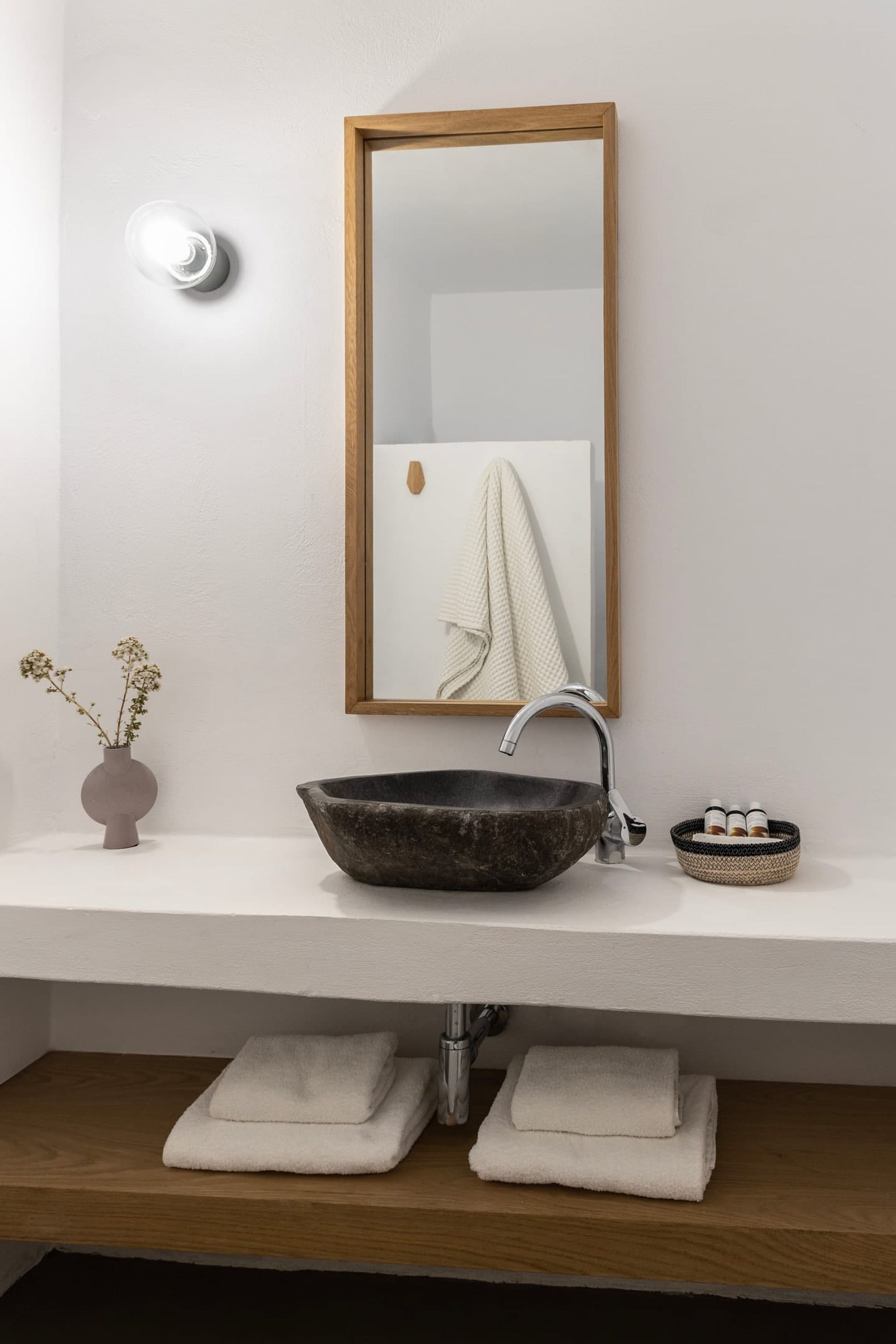 Une salle de bain avec un miroir encadré en bois, un lavabo en pierre sombre et un petit vase contenant des fleurs séchées.