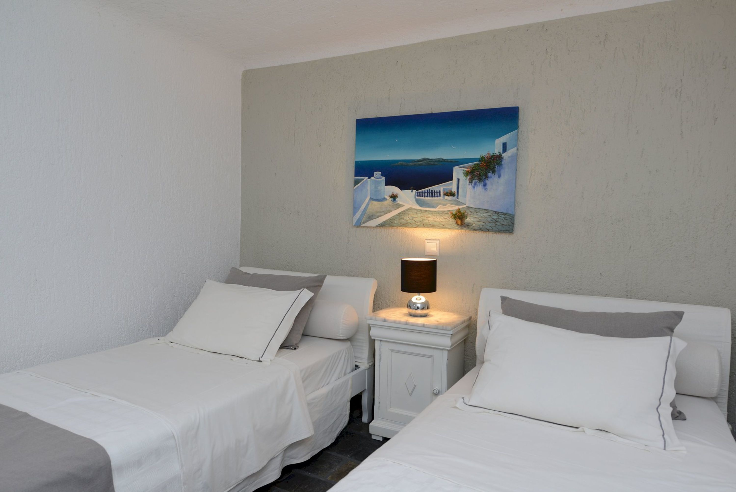 A hotel room with two single beds, a nightstand with a lamp, and a painting of a coastal scene on the wall.