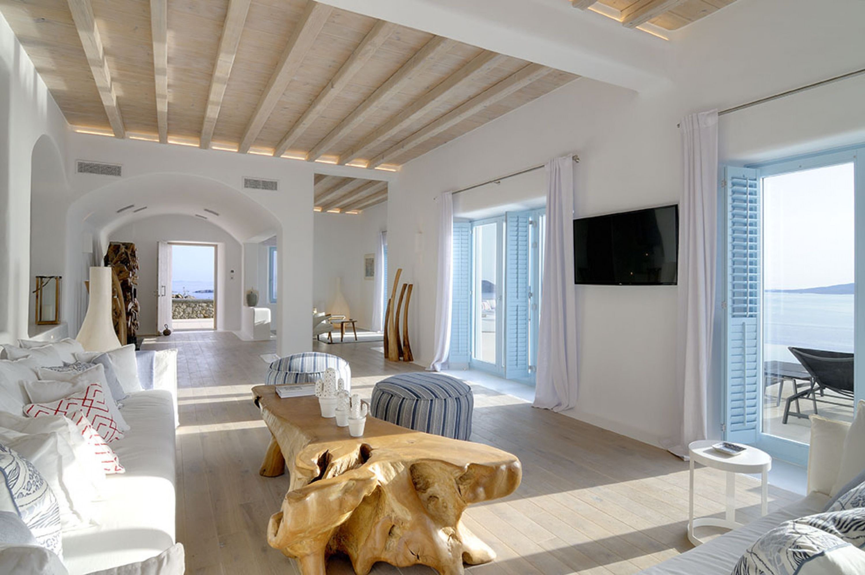 A spacious living room with a wooden coffee table, a sofa, and a TV mounted on the wall. There is a large window with shutters and a view of the sea.