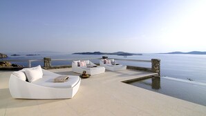 A seaside terrace with white cushioned seating and a stone balustrade overlooking calm waters. - Bluebird Villas – Villa Sunbird (Agios Ioannis Diakoftis)