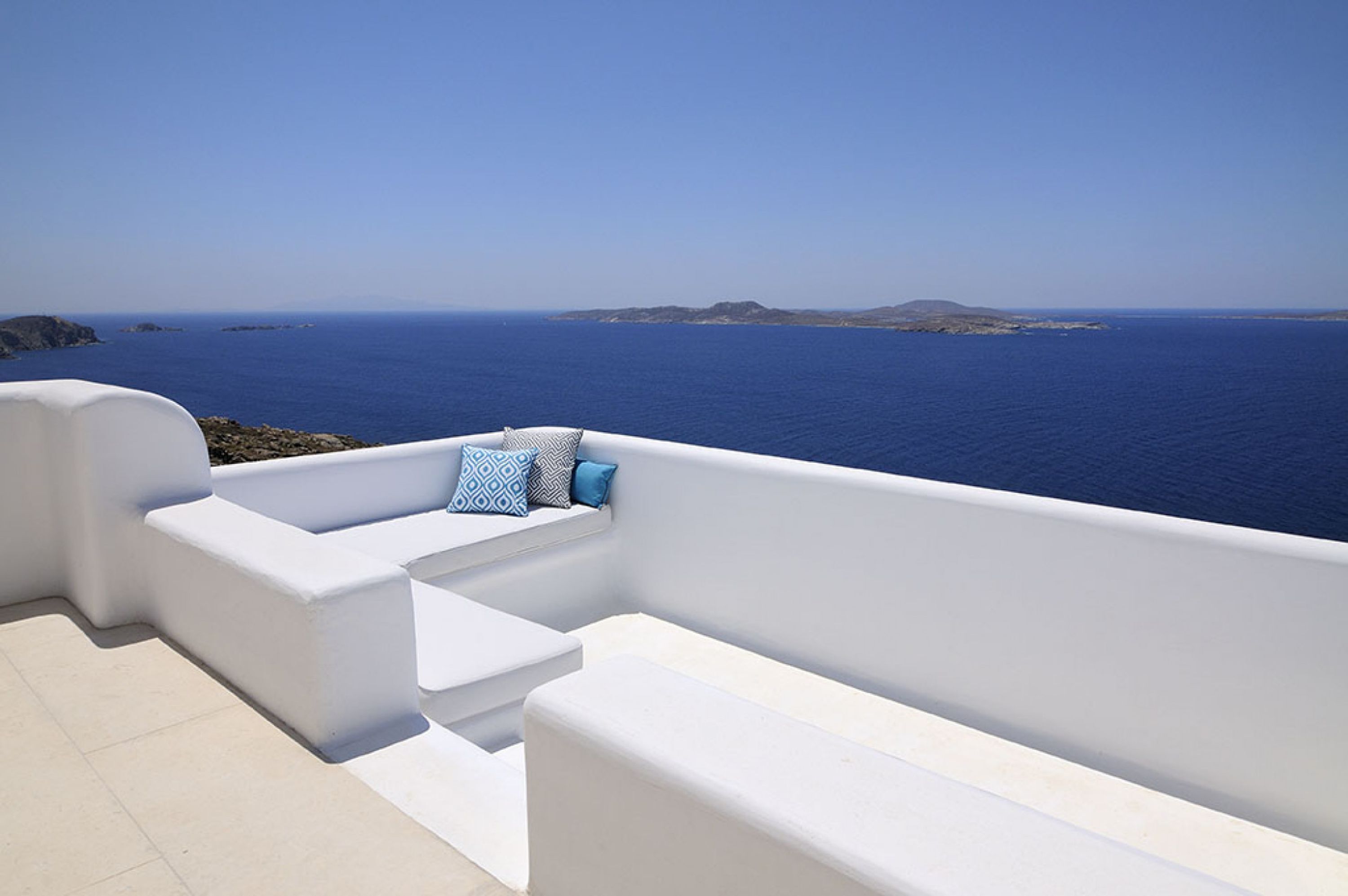 A white outdoor seating area with a view of the sea and distant islands.