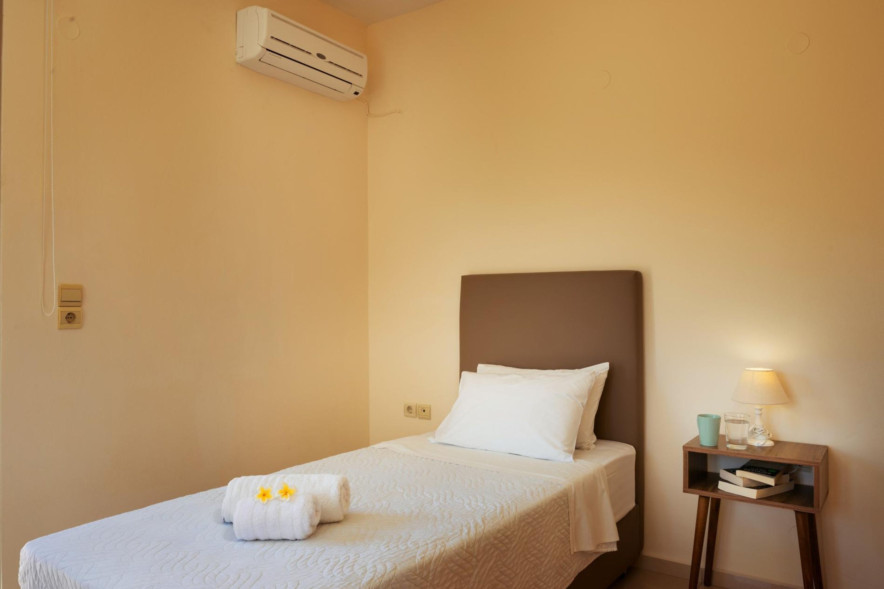 Een enkel bed met een wit hoofdeinde en kussens, een nachtkastje met een lamp, en een airconditioning op de muur.