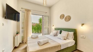 A bedroom with a green upholstered headboard, a large bed with white linens, a flat-screen TV mounted on the wall, and a sliding glass door leading to a balcony with a view of greenery. - Villa Agricola with private swimming pool (Zakynthos)