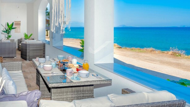 A modern room with a view of the sea, wicker furniture, and a breakfast setup on a glass table. - Sky Beachfront Villa with private pool (Prinos)