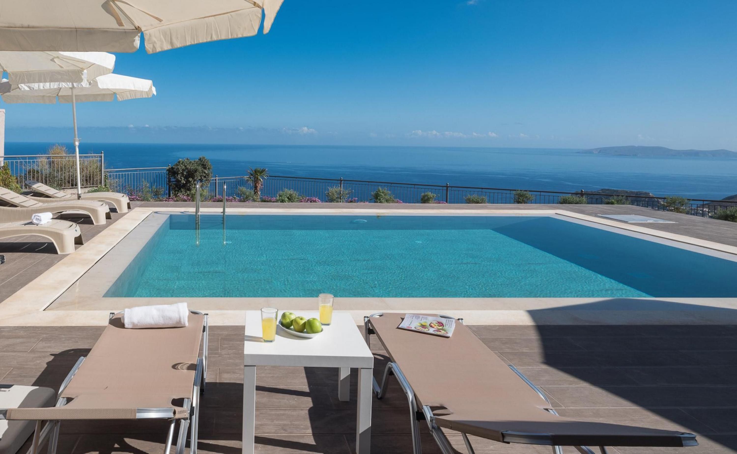 Piscina com espreguiçadeiras e guarda-sóis, mesa com bebida e frutas, e vista para o mar.