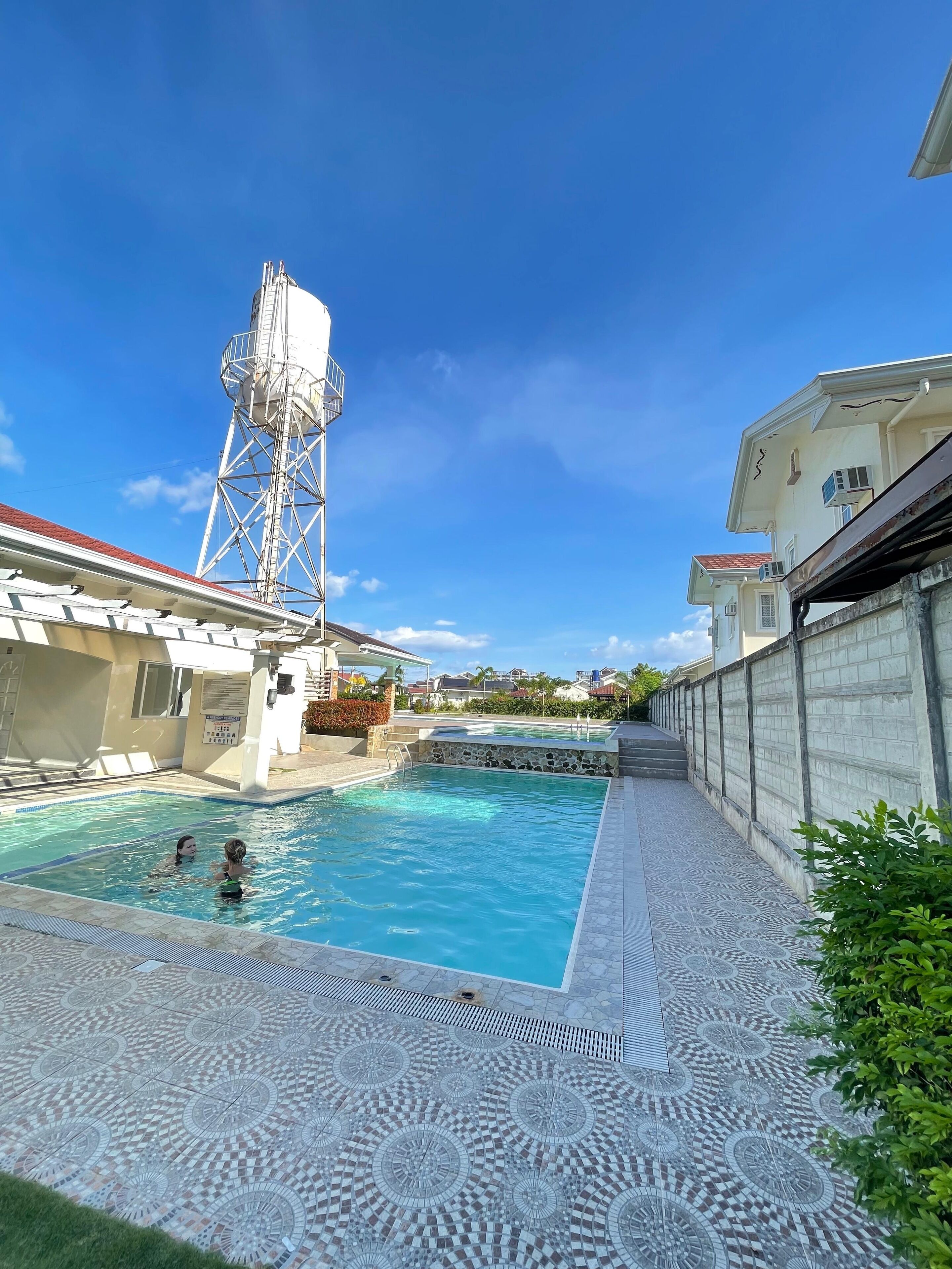 A swimming pool with two people, a tall structure, and a building with a red roof.