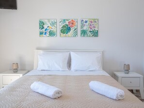 Ein ordentlich bezogenes Bett mit weißen Laken und zwei aufgerollten Handtüchern, begleitet von weißen Nachttischen mit Lampen. Darüber hängen drei gerahmte Blumenbilder an der Wand.