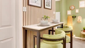 Comedor con mesa blanca para dos, sillas con cojines verdes y pared de listones de madera.