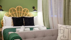 Una cama bien hecha con cubrecama verde, dos almohadas blancas y dos almohadas negras. Hay un sofá gris con una manta verde y una almohada blanca con diseño negro. Se ve un cabecero de madera con motivos de hojas y dos lámparas de pared.