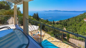 Un balcón con mesa y sillas de vidrio, con vistas a un paisaje costero con árboles y un mar azul cristalino.