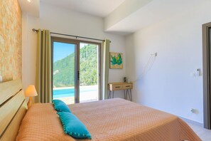 A bedroom with a bed, a nightstand, a lamp, a painting, and a view of the outdoors. - Vista Mare Assinia in Kentroma (Kentroma)