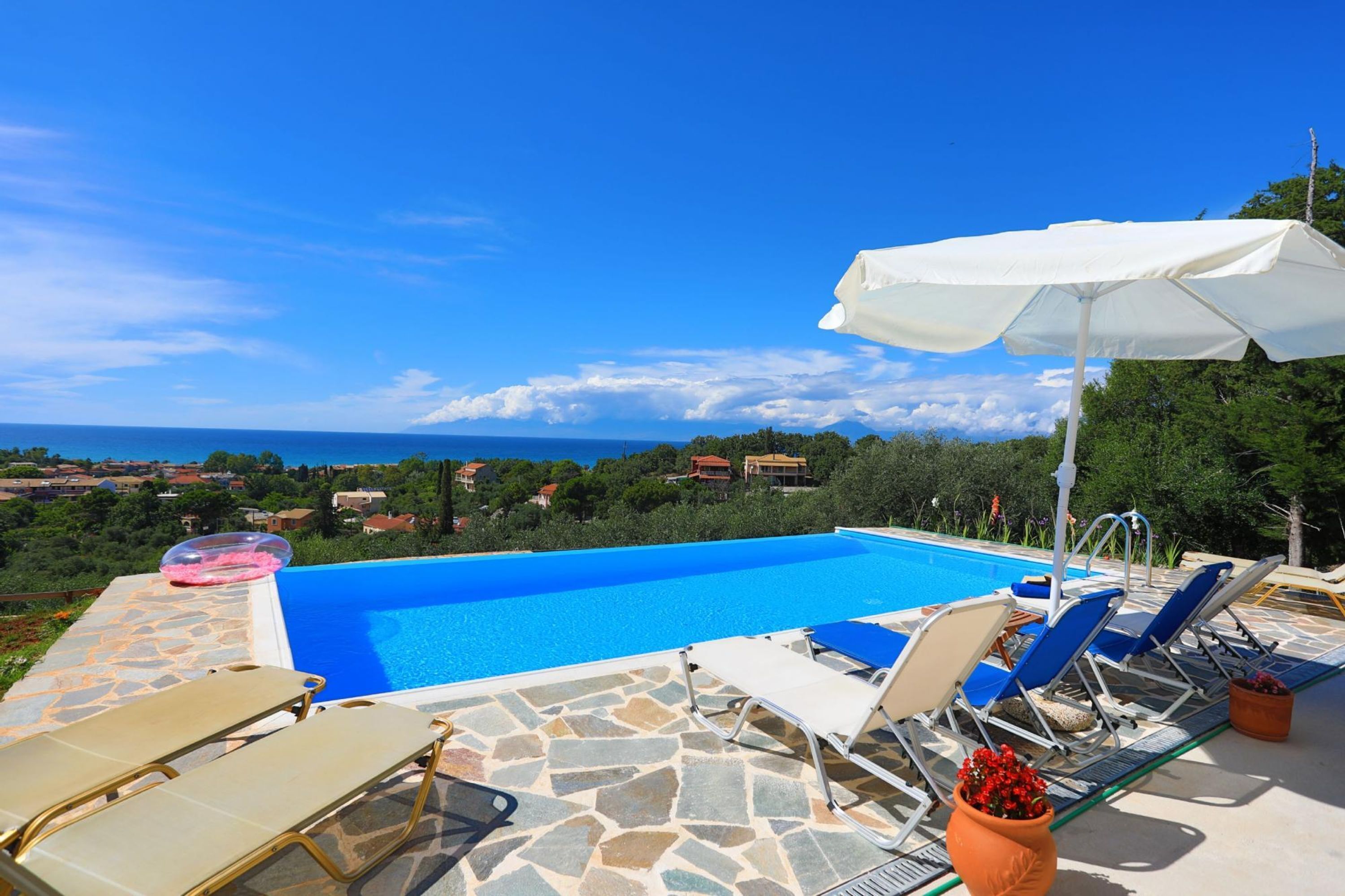 A pool area with lounge chairs and an umbrella, surrounded by stone tiles and a view of the sea.