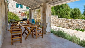 Une terrasse extérieure avec une table et des chaises en bois, des murs en pierre et une vue sur un bâtiment en pierre doté de volets verts.