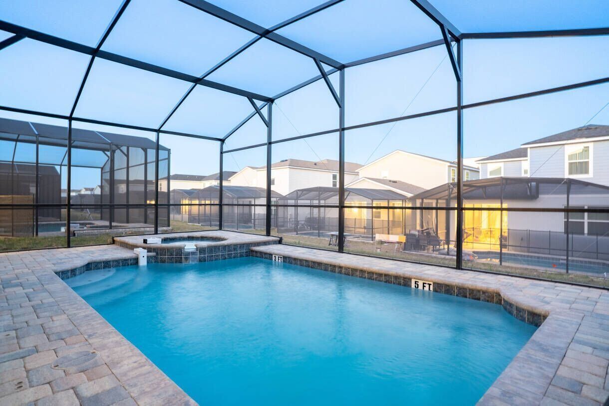 Una piscina con cerramiento de vidrio, rodeada por un patio de ladrillo y vistas a edificios residenciales.