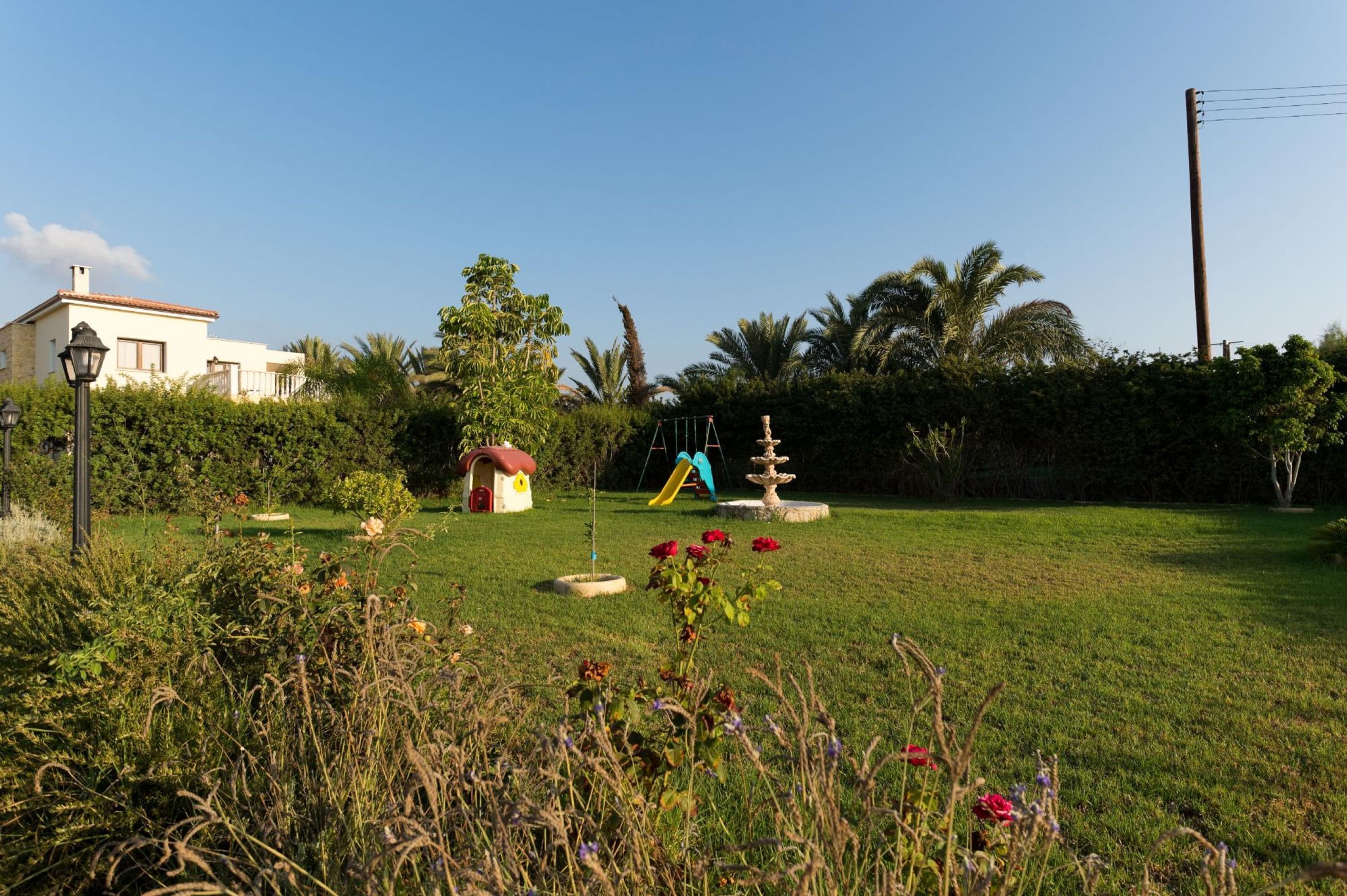 A well-maintained garden with a small play area, including a slide and a fountain, surrounded by lush greenery and a house in the background.