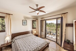 A bedroom with a bed, a ceiling fan, a nightstand, a window with curtains, and a balcony with a table and chairs. - Villa Tsikkos Tria in Argaka (Argaka)
