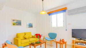 Ein Wohnzimmer mit gelber Couch, blauen Stühlen, einem Holztisch mit Blumenvase, einem Fernseher auf einem Holzschrank und einem Fenster mit blauen Jalousien.