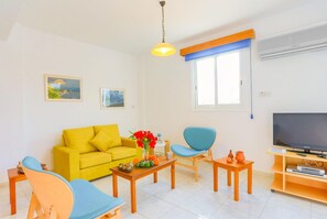 Ein Wohnzimmer mit gelber Couch, blauen Stühlen, einem Holztisch mit Blumenvase, einem Fernseher auf einem Holzschrank und einem Fenster mit blauen Jalousien.