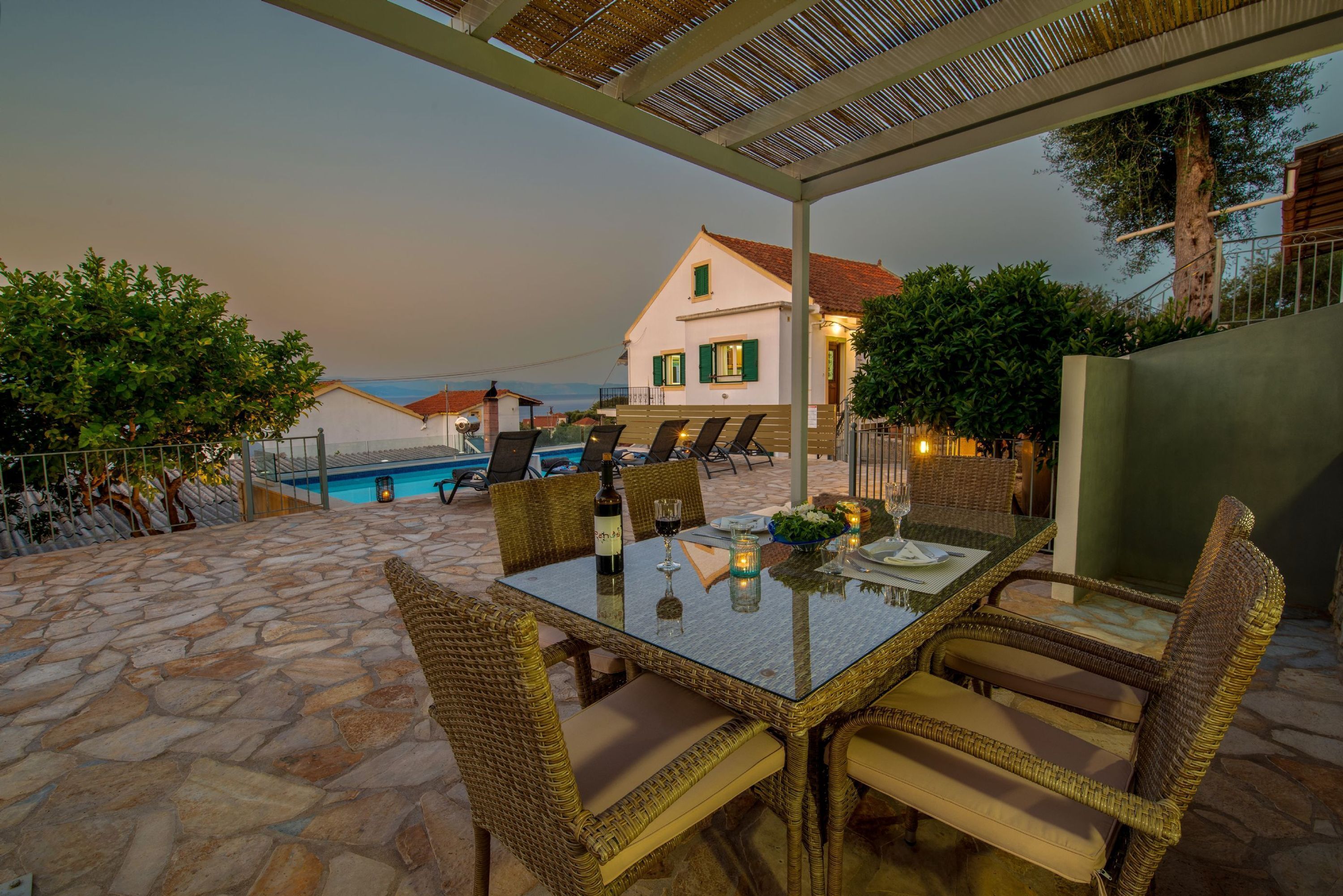 Un patio exterior con suelo de piedra, mobiliario de mimbre, una mesa de vidrio y vista a una piscina y una casa al fondo.