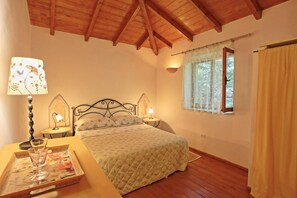 Ein Schlafzimmer mit Holzbalkendecke, einem Bett mit floralem Überwurf, zwei Nachttischlampen, einem Fenster mit Vorhängen und einem Tablett mit Glas und Buch auf dem Nachttisch.