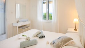 A hotel room with a bed, a bedside table with a lamp, a window with curtains, a vanity with a mirror, and a view of greenery outside. - Antigoni Villa Ena in Longos (Longos)