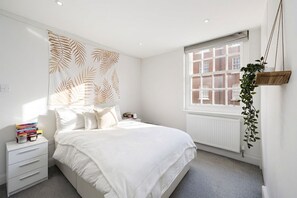 Ein Schlafzimmer mit einem Bett, einem Nachttisch mit Büchern, einem Heizkörper und einem Fenster mit Blick auf Gebäude.