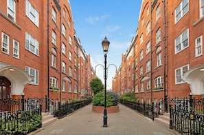 A street with red brick buildings, a central lamp post, and a circular planter with greenery. - [Superhost] 12 mins walk to Buckingham Palace & Big Ben - Cozy 2BR Family Rental (London)
