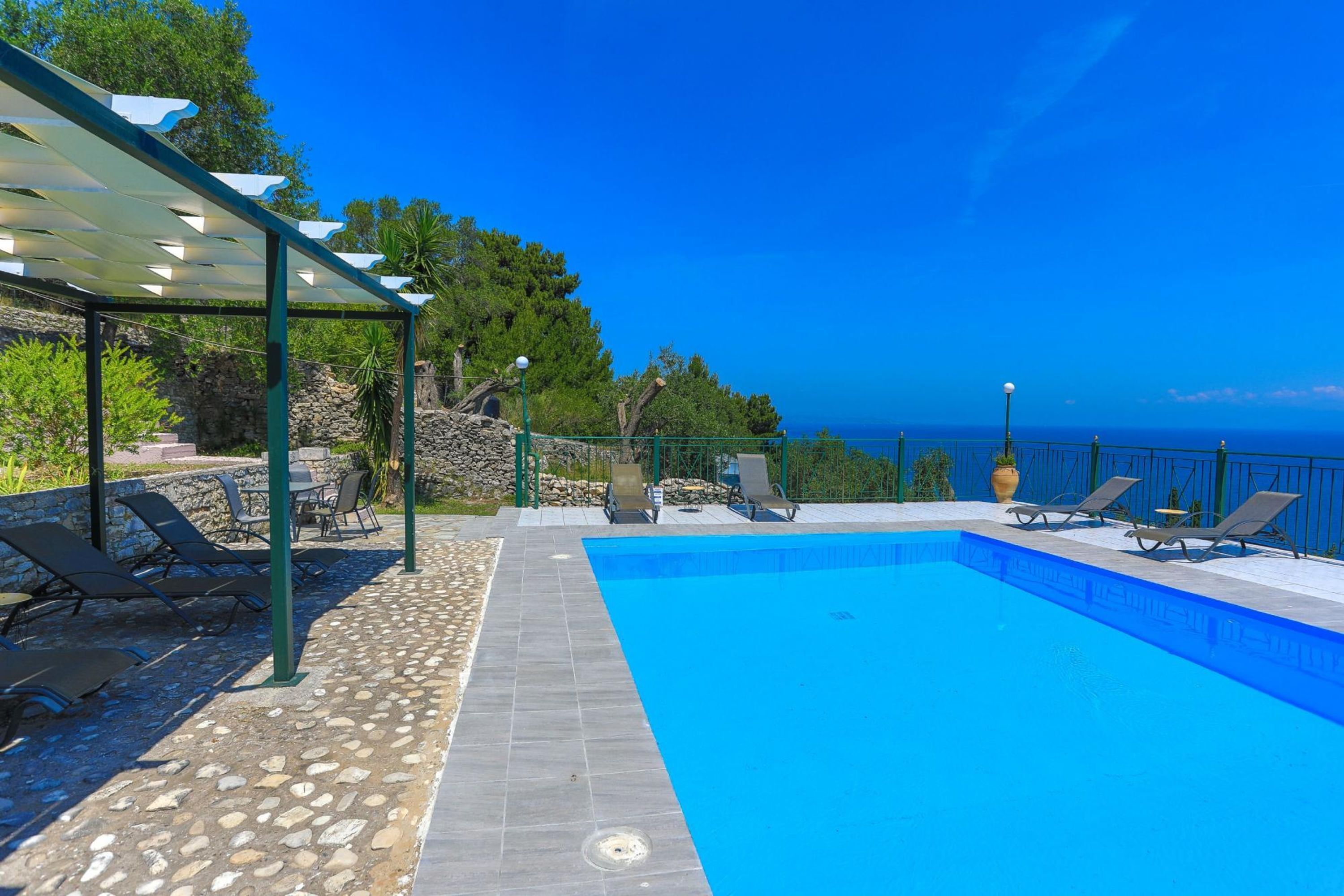 A swimming pool with a pergola and lounge chairs, surrounded by stone walls and vegetation, overlooking a clear blue sky and ocean.