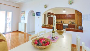 A dining area with a wooden table, chairs, and a fruit bowl. There is a kitchen visible through an archway with wooden cabinets and a refrigerator. - Villa Calypso in Nisaki (Nisaki)