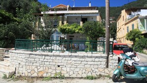 A stone wall with a green metal railing, a parked scooter, and a red car in the background. - Chrissa in Katavolos (Katavolos)
