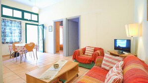 Een woonkamer met een bank, een televisie, een eettafel met stoelen, en een keuken die zichtbaar is door een open deur.