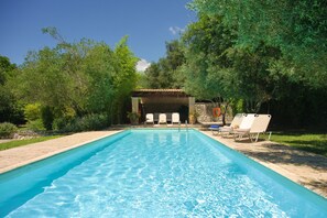 Una piscina rectangular con agua azul cristalina, rodeada por un deck de piedra. Hay sillas reclinables blancas y una zona de estar cubierta con un muro de piedra.