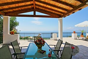 Eine Terrasss mit Holz-Pergola, einem Glastisch mit Stühlen, einem Blumenstrauß und Blick aufs Meer.