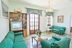 A living room with green patterned sofas, a wooden bookshelf, a television, and a chandelier. - Villa Alexandros in Nisaki (Katavolos)