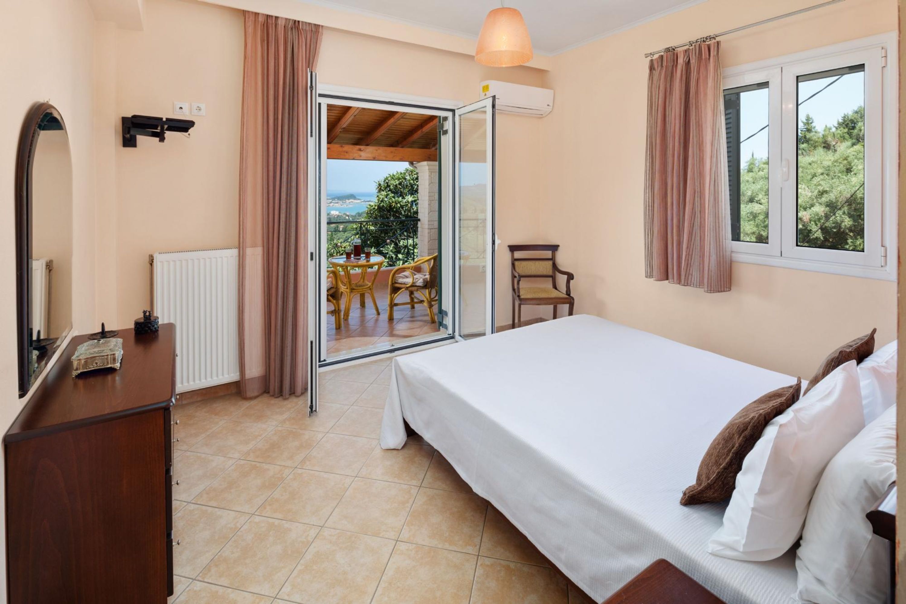 Une chambre avec un lit, une commode, une chaise et une vue sur un espace extérieur aménagé avec une table et des chaises, accessible par une porte ouverte.