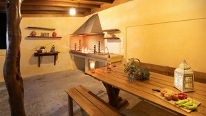 An outdoor dining area with a wooden table, benches, and a lantern. There is a tree trunk to the left and a kitchen area in the background. - Sunset House in Sidari (Corfu)