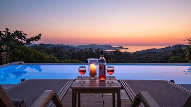A wooden table with a bottle and two glasses of red wine, set against a sunset view over a swimming pool. - Sunset House in Sidari (Corfu)