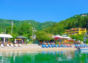 A beachside area with sun loungers, a boat, and buildings in the background. - Villa Triantafyllo in Kentroma (Kentroma)