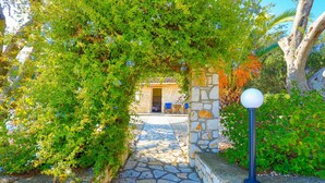 A stone pathway leading to a building with a covered entrance, surrounded by lush greenery and a stone wall. - Villa Helona in Lithiasmenos (Lithiasmenos)