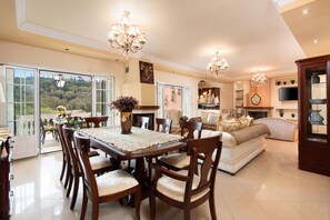 A spacious living area with a dining table, chairs, and a sofa. There are chandeliers, a fireplace, and a television mounted on the wall. - Eleonas in Kato Korakiana (Corfu)