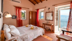 Una camera da letto con un letto, un armadio in legno, un comodino, uno specchio e vista sul mare attraverso una porta aperta.