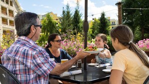 Un grupo de personas cenando al aire libre en una mesa con estructura metálica, rodeado de follaje y flores.