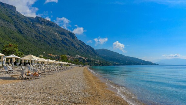 Una playa de guijarros con tumbonas y sombrillas, un mar azul cristalino y montañas al fondo.