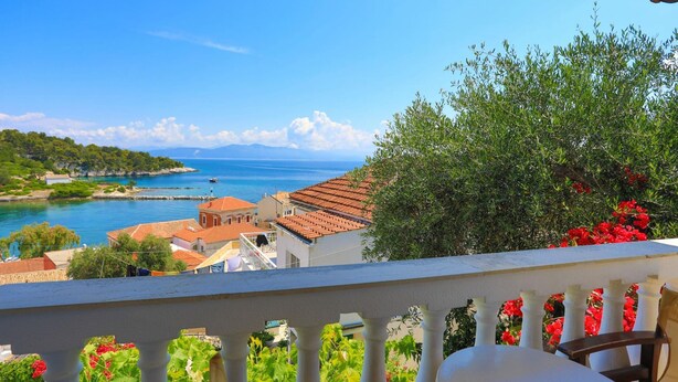 Ein Balkon mit Blick auf eine Küstenstadt, darunter Häuser mit roten Dächern und ein klares, blaues Meer.