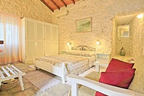 A bedroom with a stone wall, a bed with a floral bedspread, a nightstand, a red chair, and a white wardrobe. - Korinas Cottage in Kentroma (Kentroma)