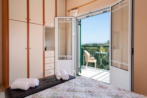 A bedroom with a bed, a bench, a wardrobe, and a sliding glass door leading to a balcony with a table and chair. - Villa Yiotta in Paxi (Paxi)