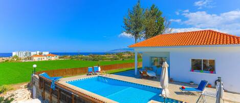 A swimming pool with lounge chairs and umbrellas, surrounded by a wooden fence and a view of the sea.