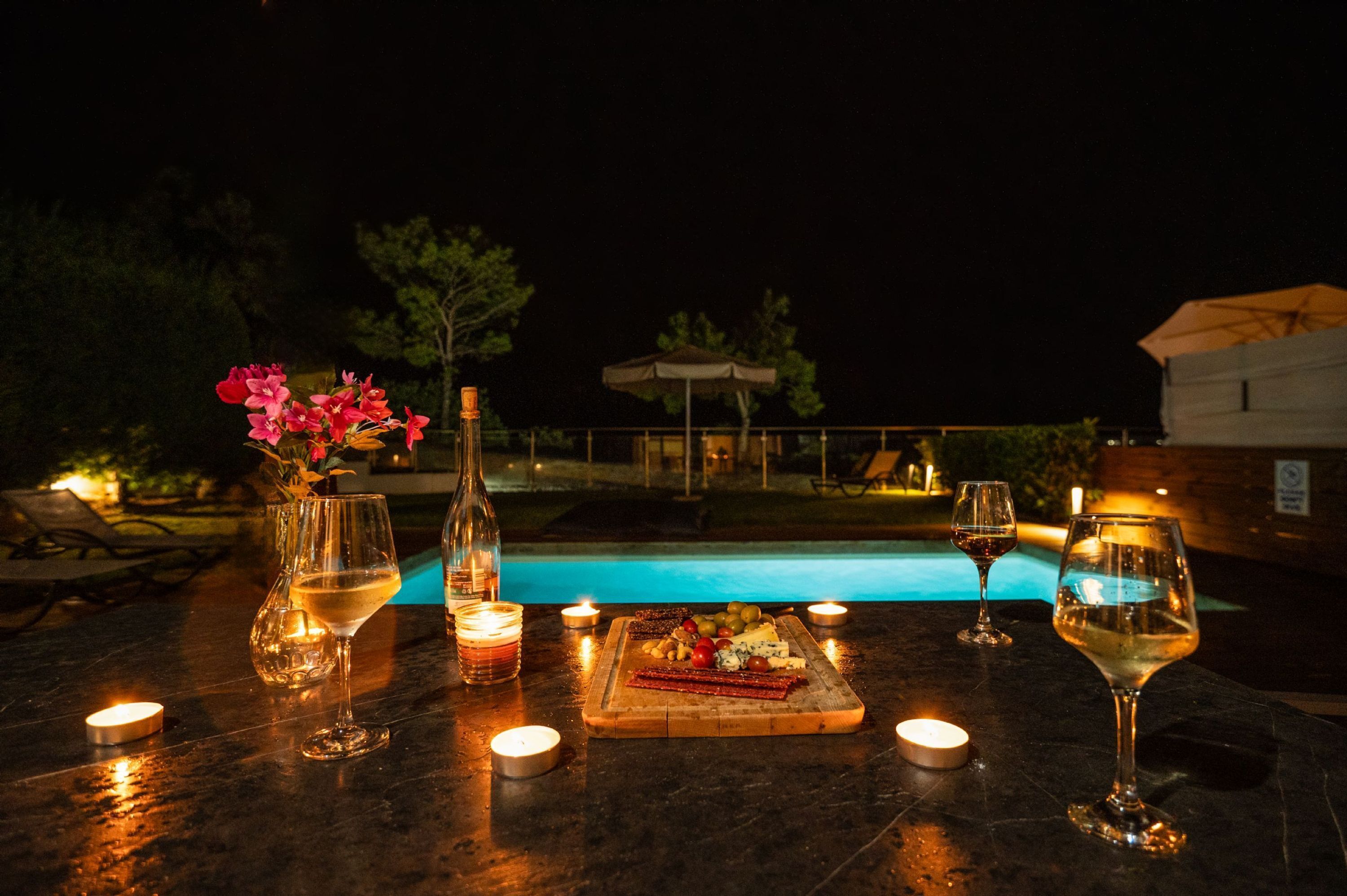 Une table dressée avec des verres à vin, une bouteille, des bougies et une planche de charcuterie, le tout placé au bord d’une piscine, la nuit.