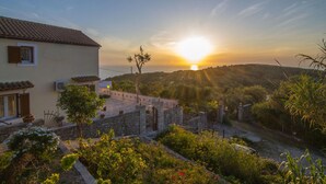 Una villa con un muro de piedra, un jardín con diversas plantas y vistas al atardecer sobre un paisaje montañoso.