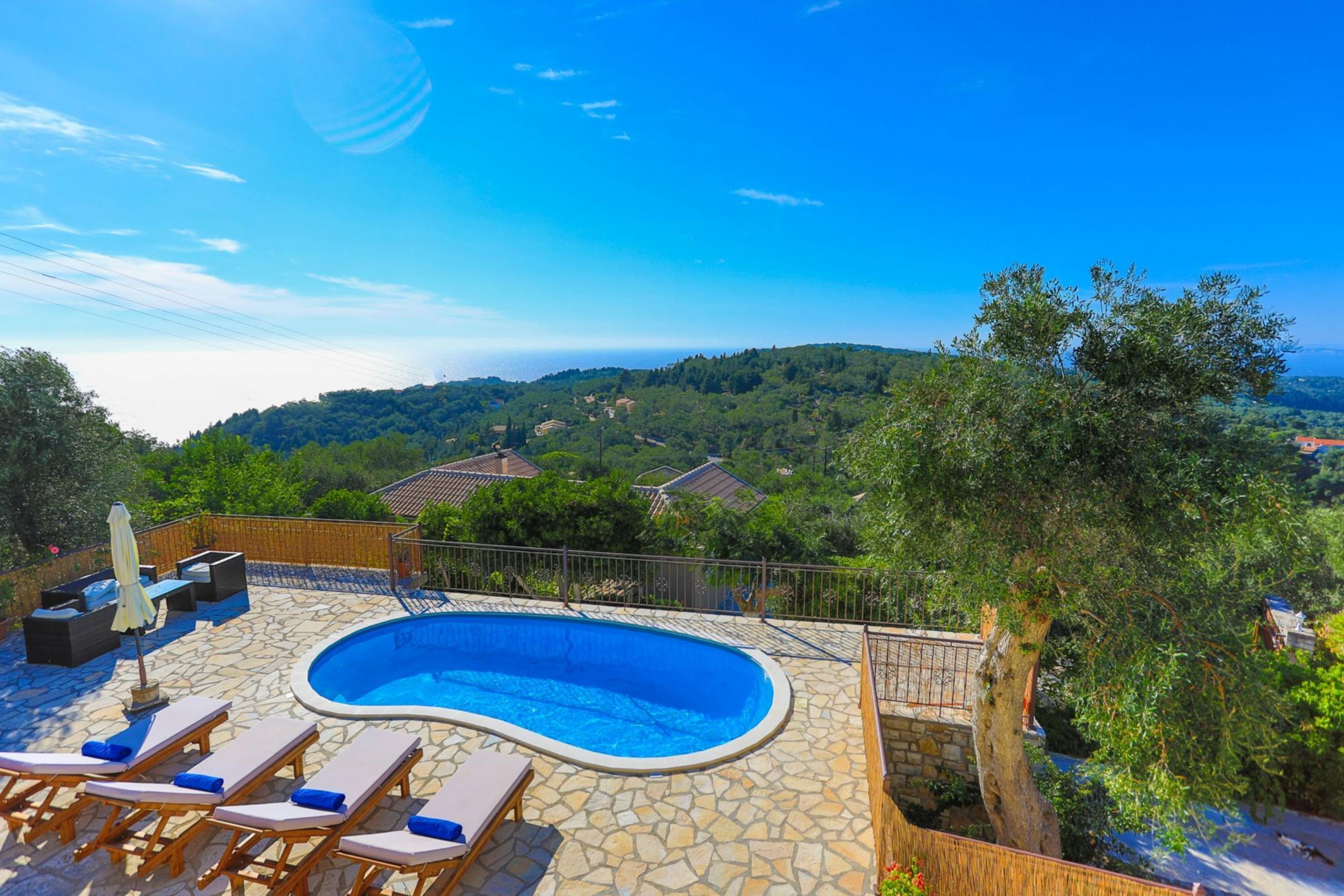 A swimming pool with lounge chairs, surrounded by stone flooring and a wooden fence, overlooking a scenic landscape with hills and houses.